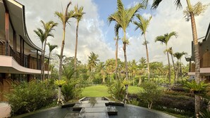 A wet pathway leading to a building with a modern design, surrounded by palm trees and a grassy area.