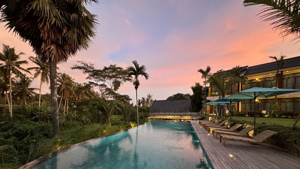A swimming pool with a wooden deck and lounge chairs, surrounded by palm trees and a building with a sloped roof.