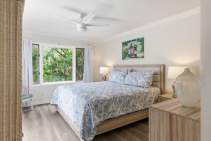 Une chambre à coucher avec un lit, deux lampes de chevet, un ventilateur de plafond, une fenêtre avec des rideaux, un fauteuil en osier et un tableau accroché au mur.