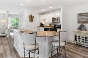 Una cocina moderna con armarios blancos, encimera de granito, electrodomésticos de acero inoxidable y una isla central con taburetes de bar.