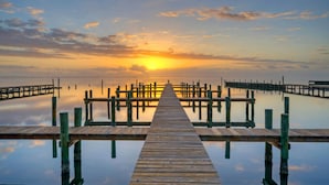 A wooden pier extending into calm waters, surrounded by dock posts, leading to a distant sunset. - 2 Waterfront Homes w/ Views, Pool, Boat Dock | Laguna Village Oasis by AvantStay (Corpus Christi)