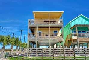 A two-story wooden house with a balcony on the upper floor, surrounded by palm trees and a fence.