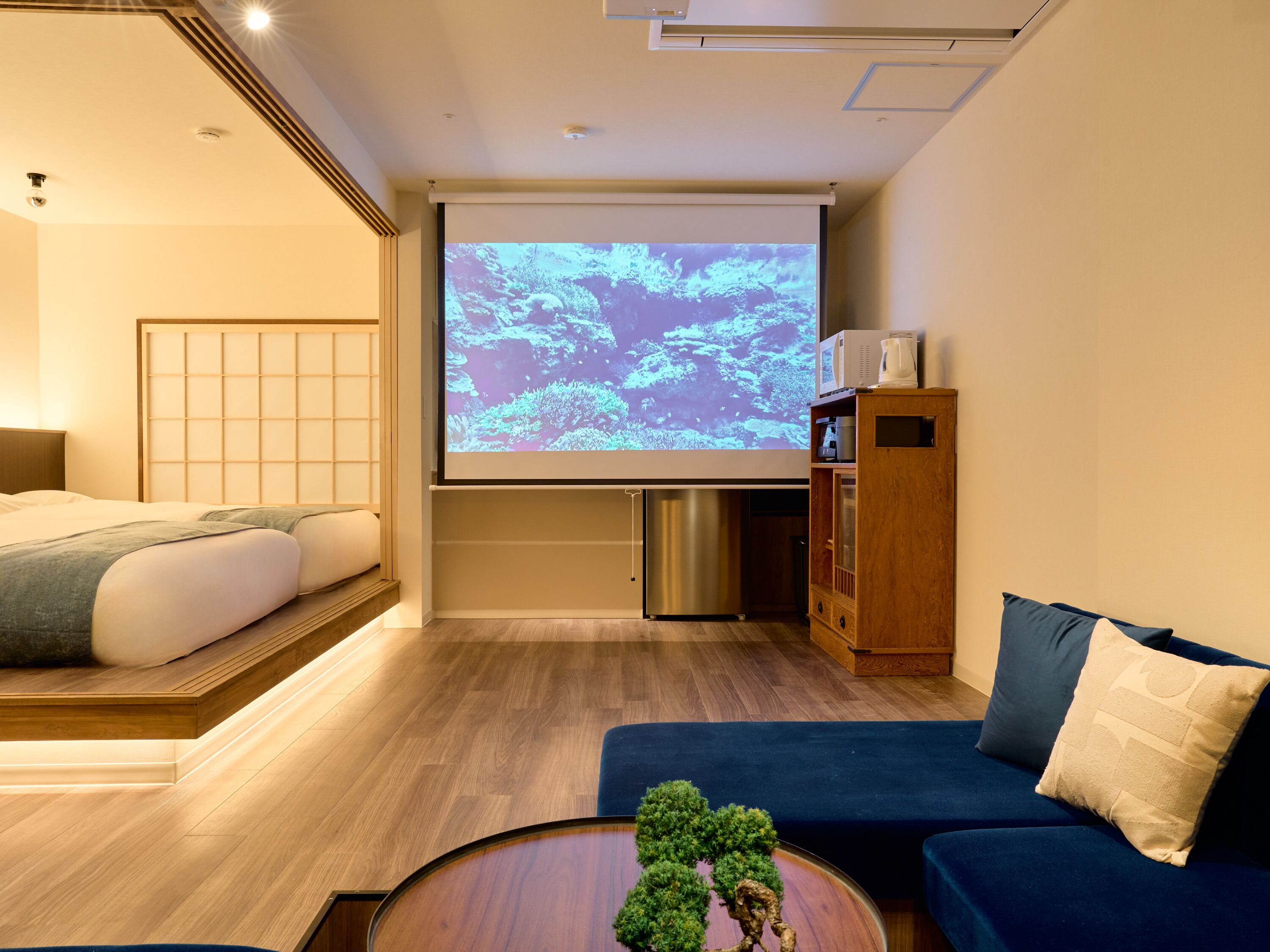 Una habitación de hotel con cama, sofá, cafetera, pantalla de proyector y un mueble pequeño.
