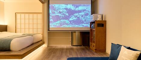 Una habitación de hotel con cama, sofá, cafetera, pantalla de proyector y un mueble pequeño.