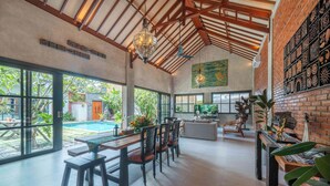 A spacious indoor area with a pool visible through large windows, a dining area with wooden furniture, and a brick wall with decorative panels. - Villa Jonsu Uluwatu by Nagisa Bali (Ungasan)