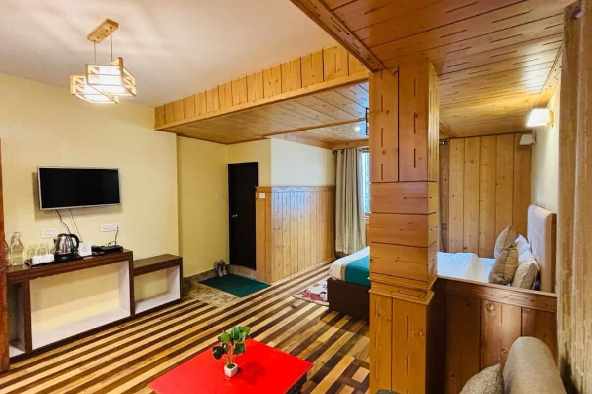 Een kamer met houten vloer, een televisie, een rode tafel met een plant, een bankje en een bed met kussens.