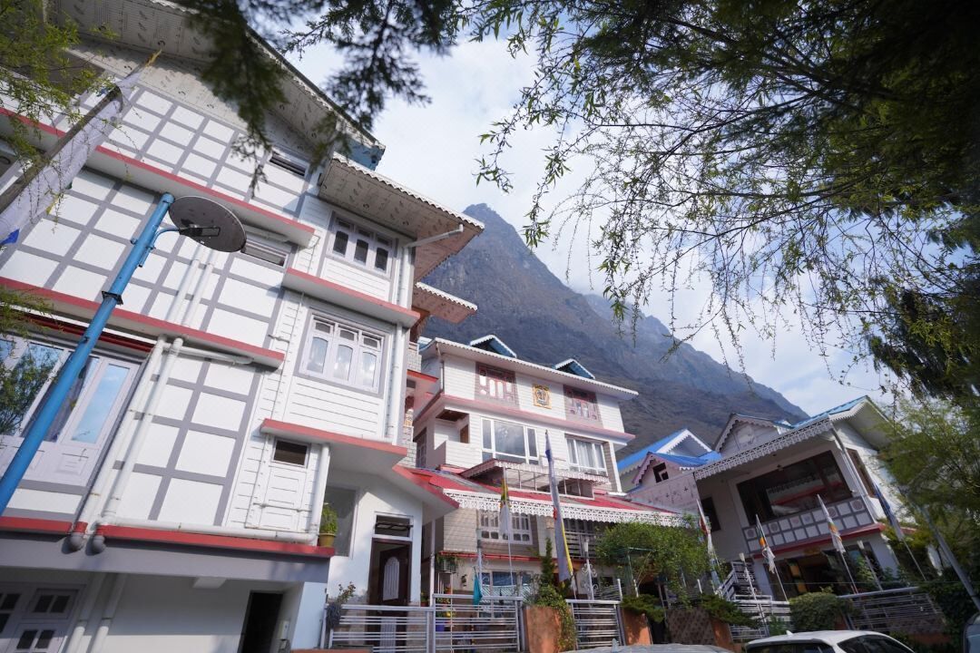 Un edificio tradicional de varios pisos con fachada roja y blanca, una antena parabólica y un paisaje montañoso de fondo.
