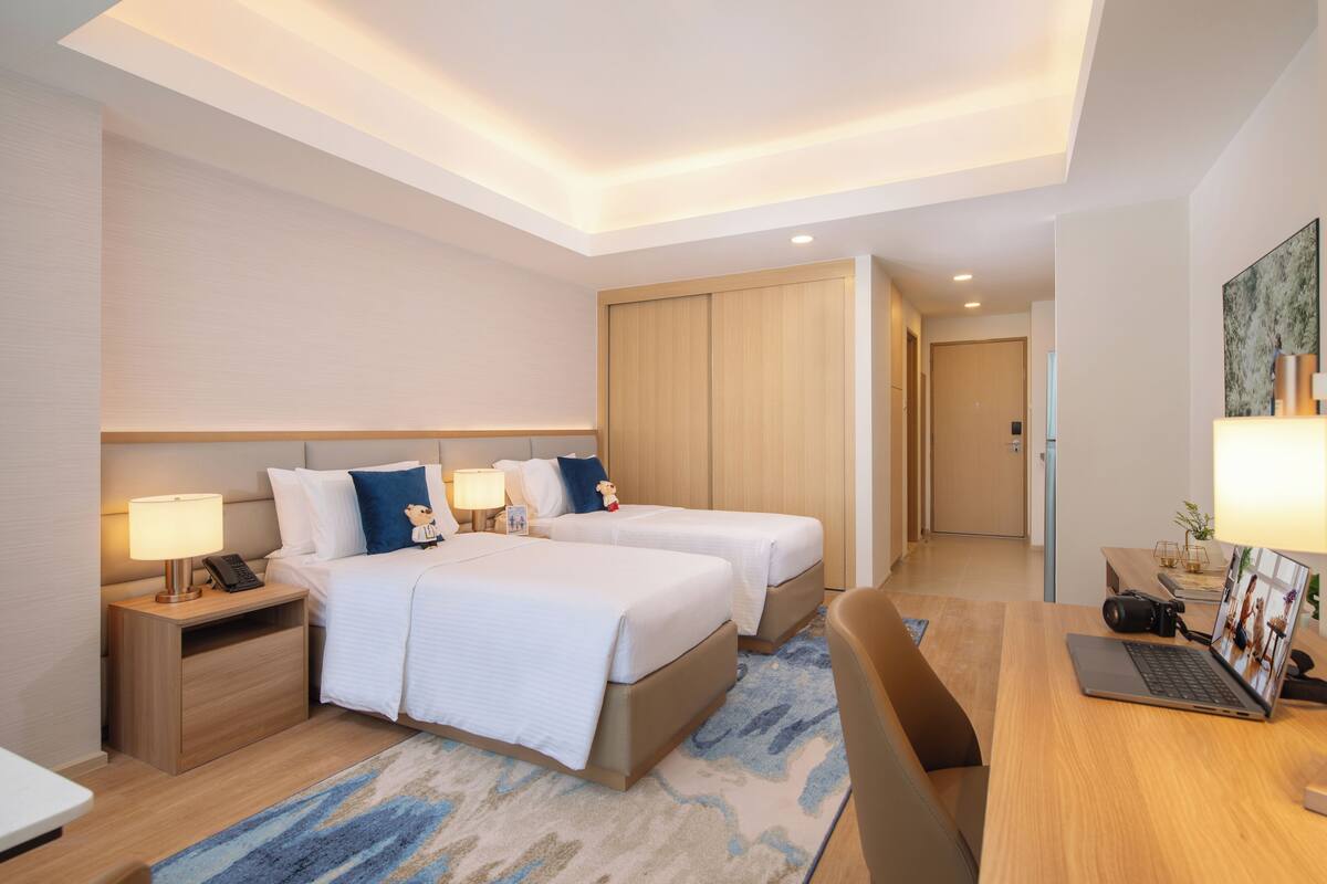 Quarto de hotel com duas camas, uma mesa de escritório com um laptop, uma câmera e uma mesa de cabeceira com abajur.