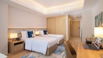 A hotel room with two beds, a desk with a laptop, a camera, and a nightstand with a lamp.