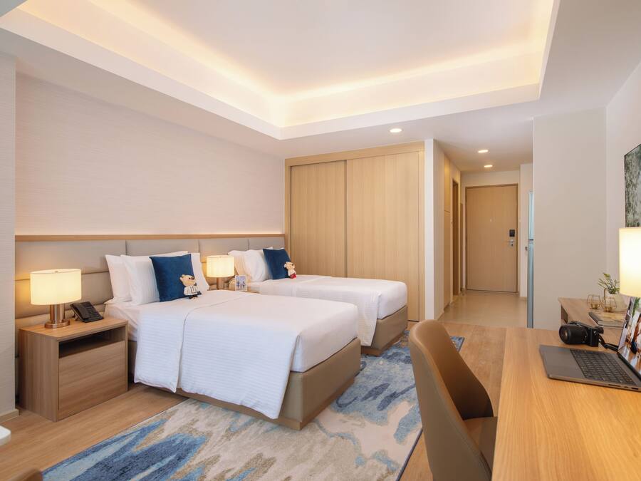 A hotel room with two beds, a desk with a laptop, a camera, and a nightstand with a lamp.
