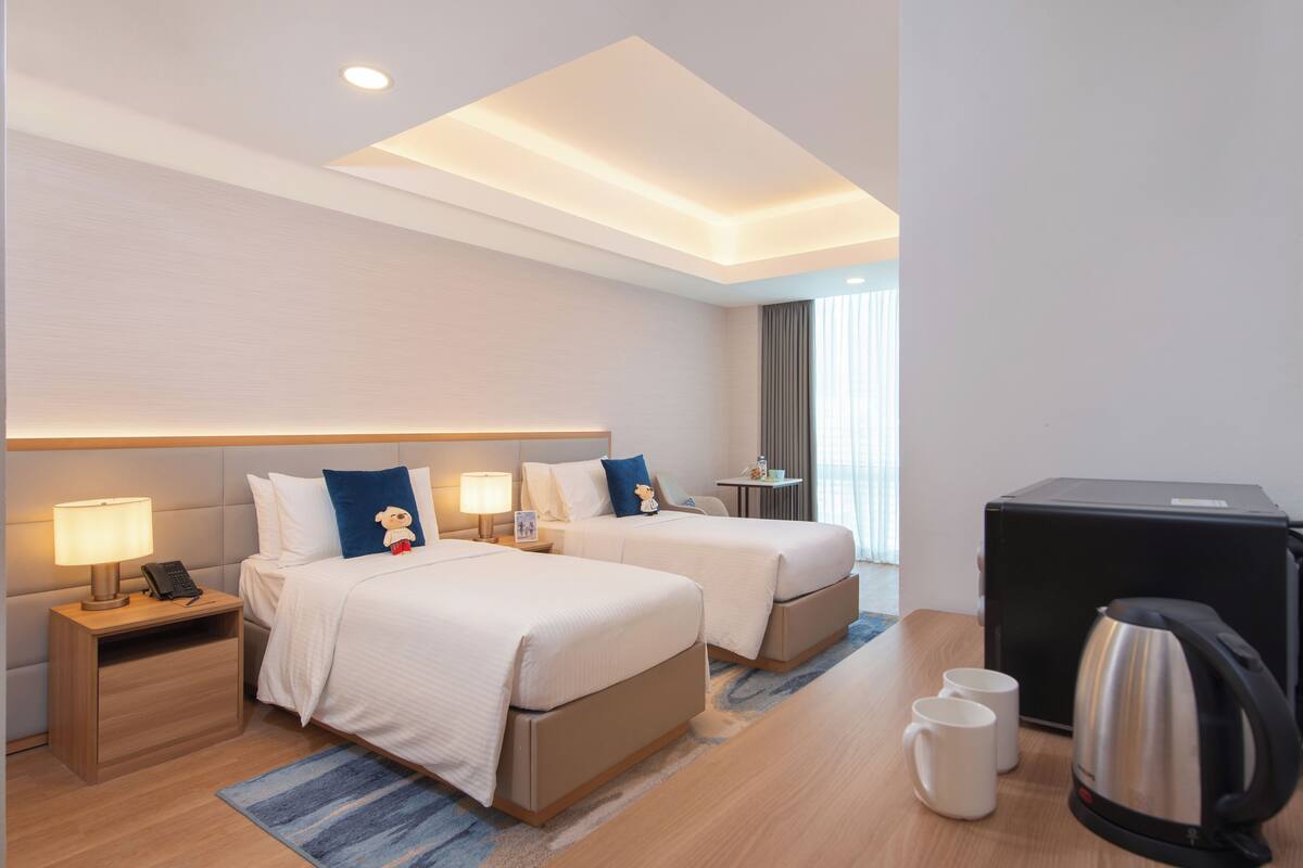 Quarto de hotel com duas camas, uma escrivaninha, uma máquina de café e uma pequena mesa com abajur.