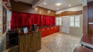Un área de recepción con un mostrador de madera, cortinas rojas, alfombra estampada y vista al exterior a través de una ventana.