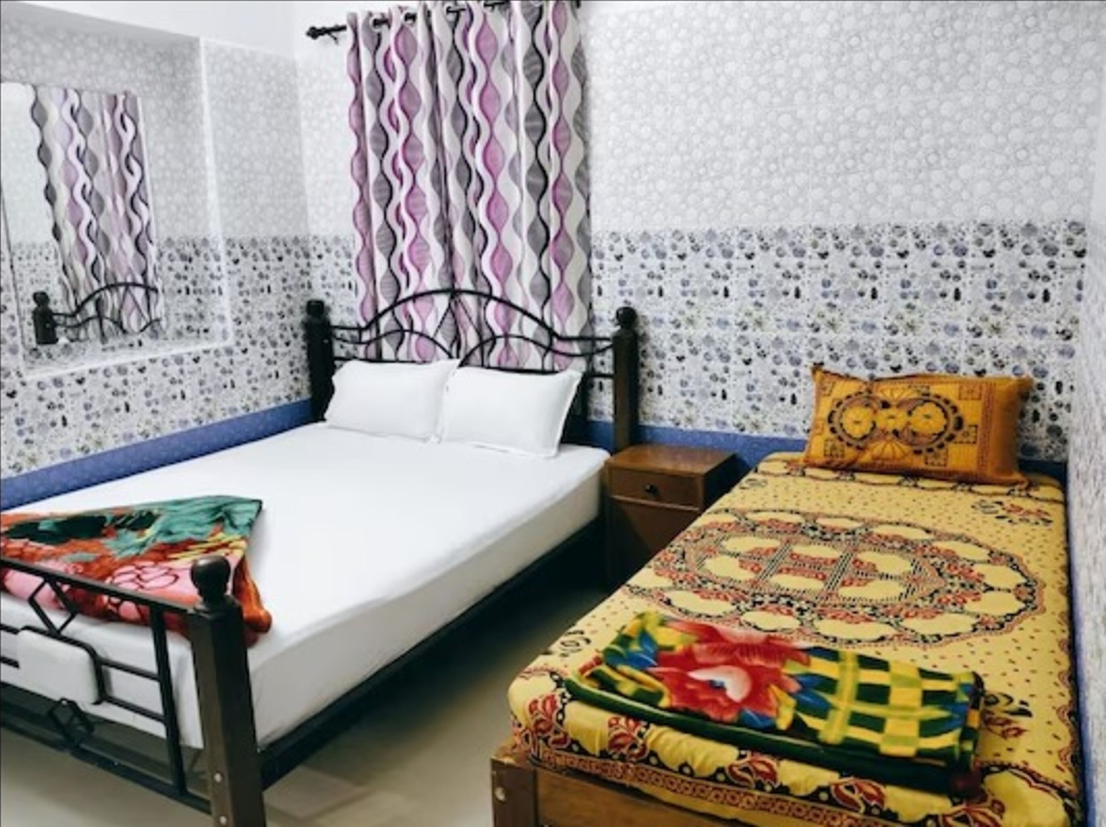 A room with two beds, one with a white bedspread and the other with a colorful bedspread. There are patterned curtains on the windows, and the walls have a mosaic tile design.