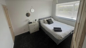 A bedroom with a bed, bedside table, lamp, and a window with blinds. - 3-bed House Sleeps 5 - Smart Tv - Free Parking (Leighton Buzzard)