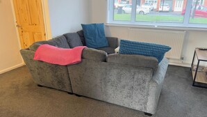 A grey sofa with a pink throw blanket and blue pillows, positioned in front of a window with a view of parked cars and buildings. - 3-bed House Sleeps 5 - Smart Tv - Free Parking (Leighton Buzzard)