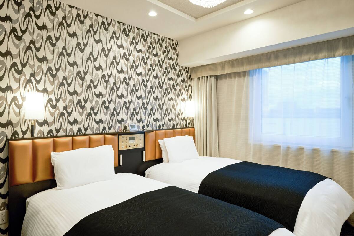 A hotel room with two beds, a control panel, a wall with a wavy pattern, and a window with curtains.