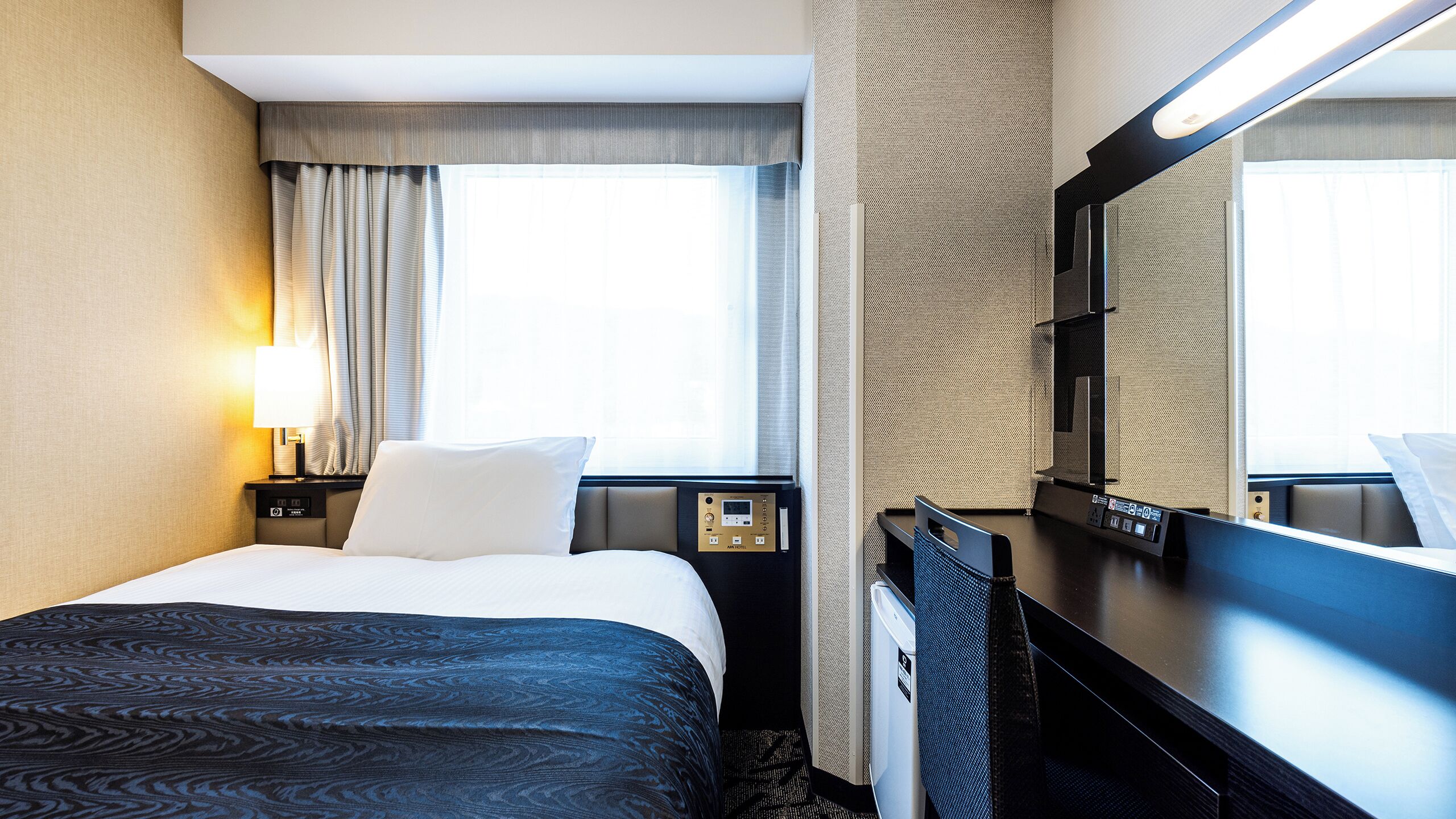 Ein Hotelzimmer mit Bett, Schreibtisch, Stuhl, einem großen Spiegel, einem Fenster mit Vorhängen und einer Bedienkonsole.