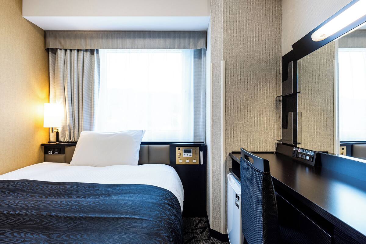 A hotel room with a bed, a desk, a chair, a large mirror, a window with curtains, and a control panel.
