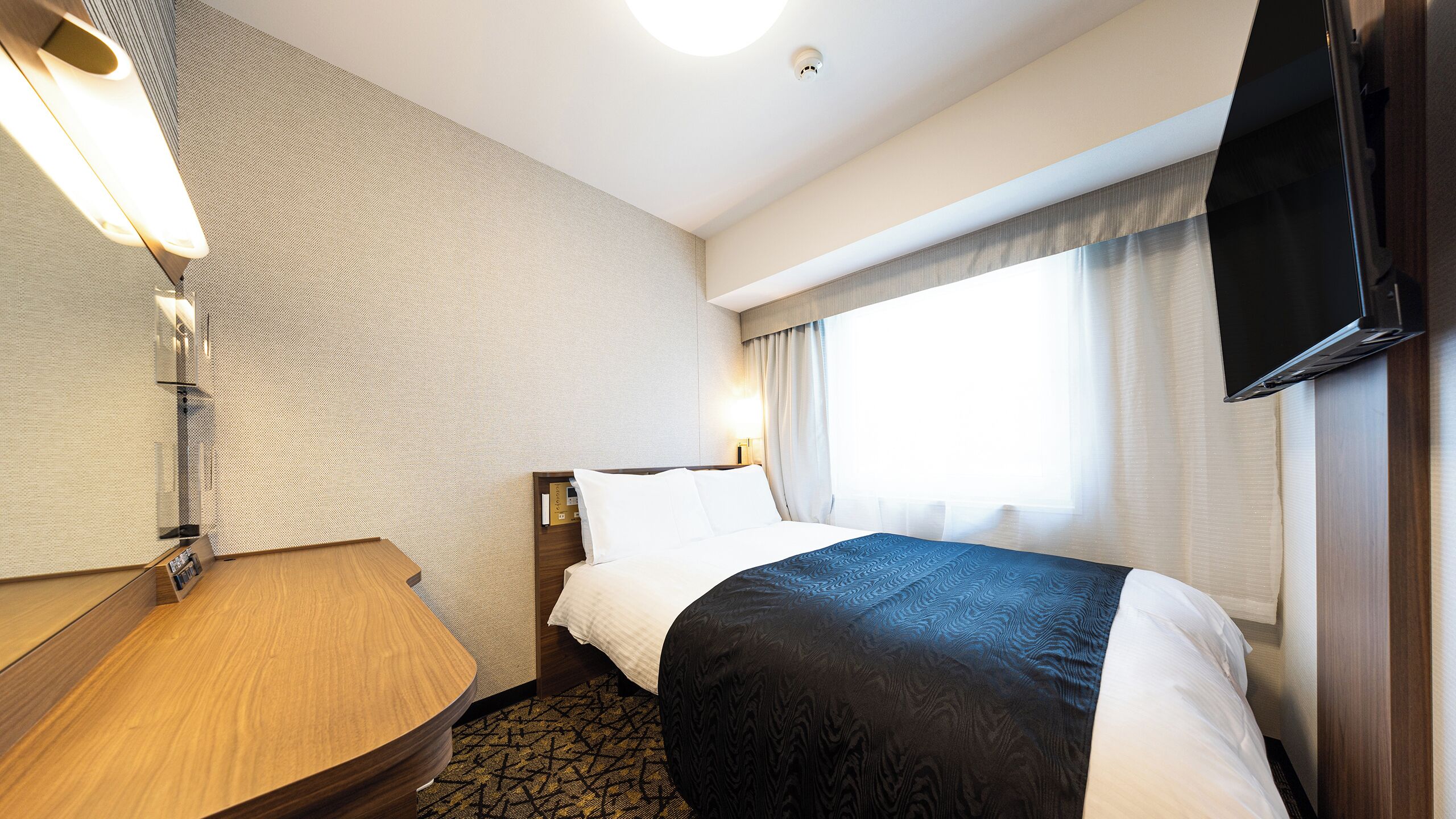 Ein Hotelzimmer mit Bett, Schreibtisch, Flachbildfernseher, einem Fenster mit Vorhängen und einer Deckenleuchte.