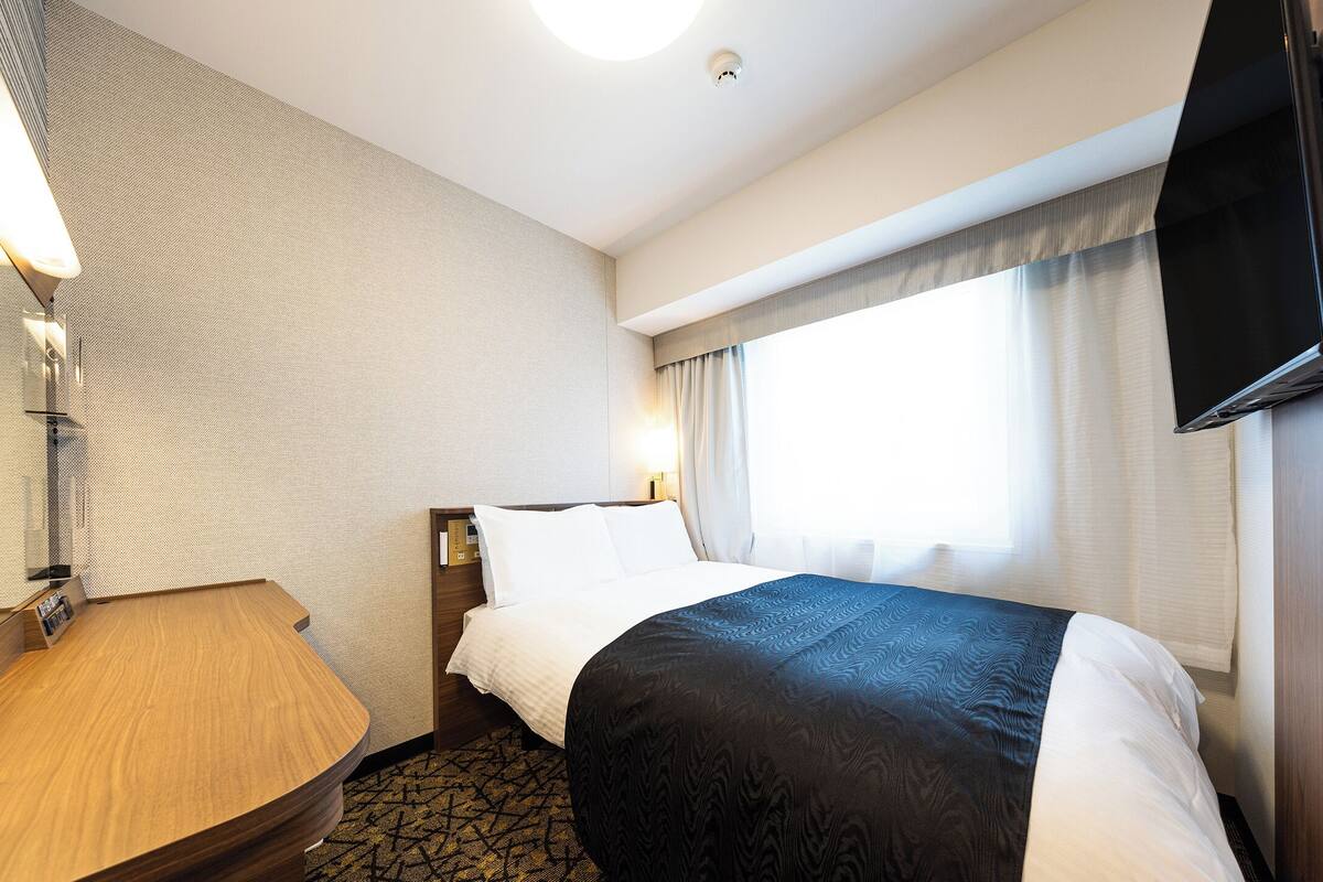 A hotel room with a bed, a desk, a flat-screen TV, a window with curtains, and a ceiling light.