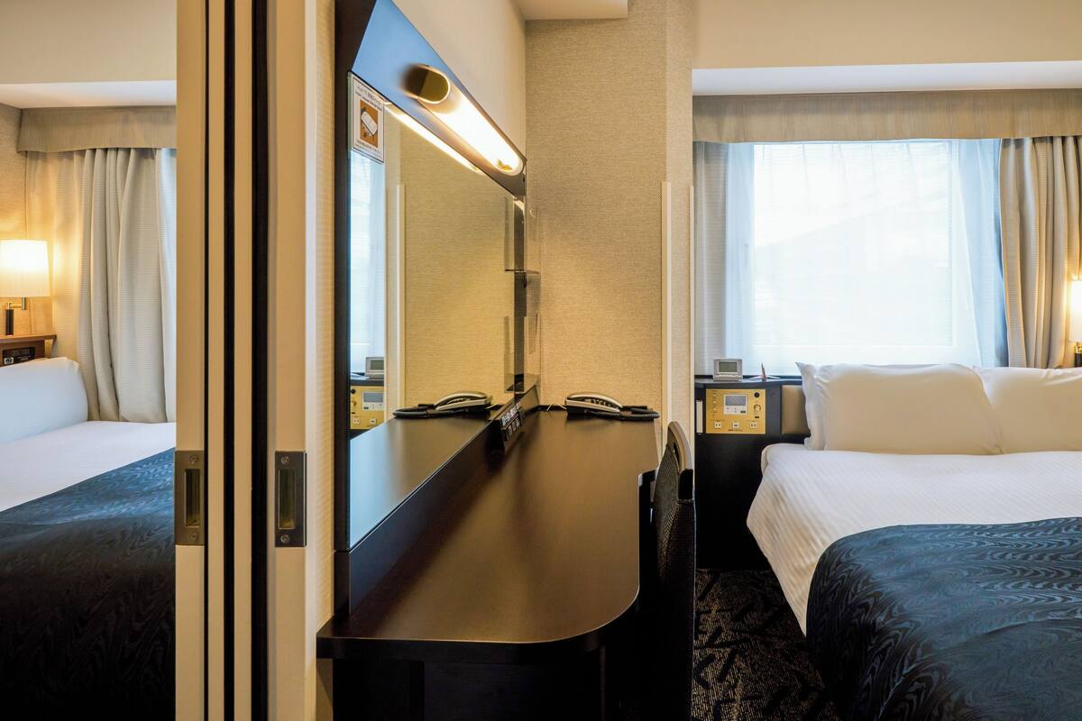 A hotel room with a bed, a desk with a telephone, a mirror, and a window with curtains.