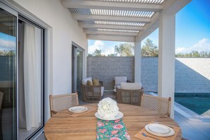 Eine Terrasse im Freien mit einem runden Holztisch, gedecktem Esstisch, Korbstühlen, einer Steinmauer und einem Pool im Hintergrund.