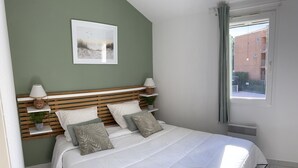 Une chambre à coucher bien rangée, avec un lit, des tables de chevet, une étagère fixée au mur, un tableau encadré et une fenêtre donnant sur des immeubles.