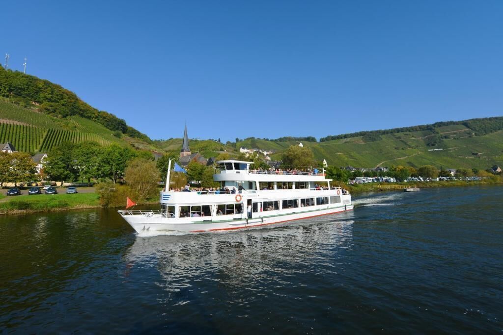 A riverboat with multiple decks, surrounded by lush green hills and a clear blue sky.
