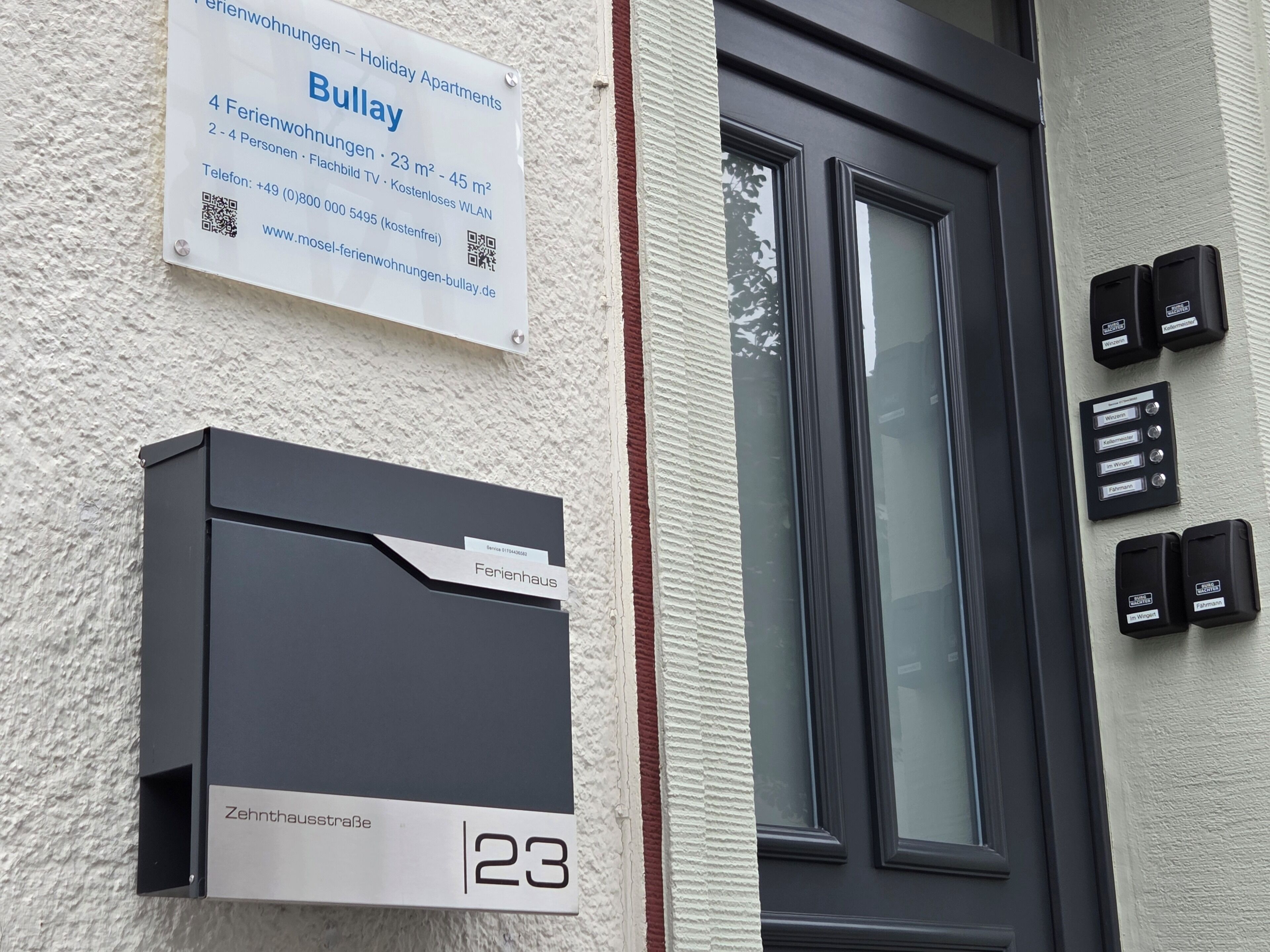 L’entrée d’un immeuble, avec une enseigne indiquant « Appartements de vacances Bullay », une boîte aux lettres et un système d’interphone.