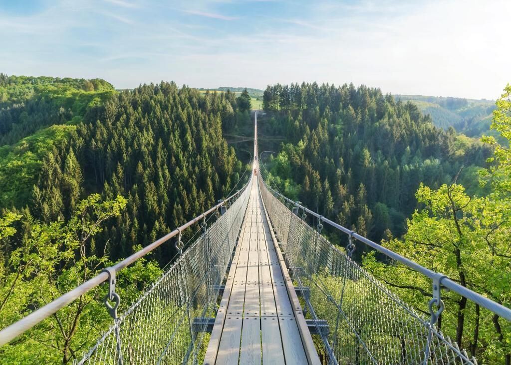 Riippusilta, jossa on puinen kävelytie ja metallikaiteet, ja sen ympärillä on tiheää metsää.