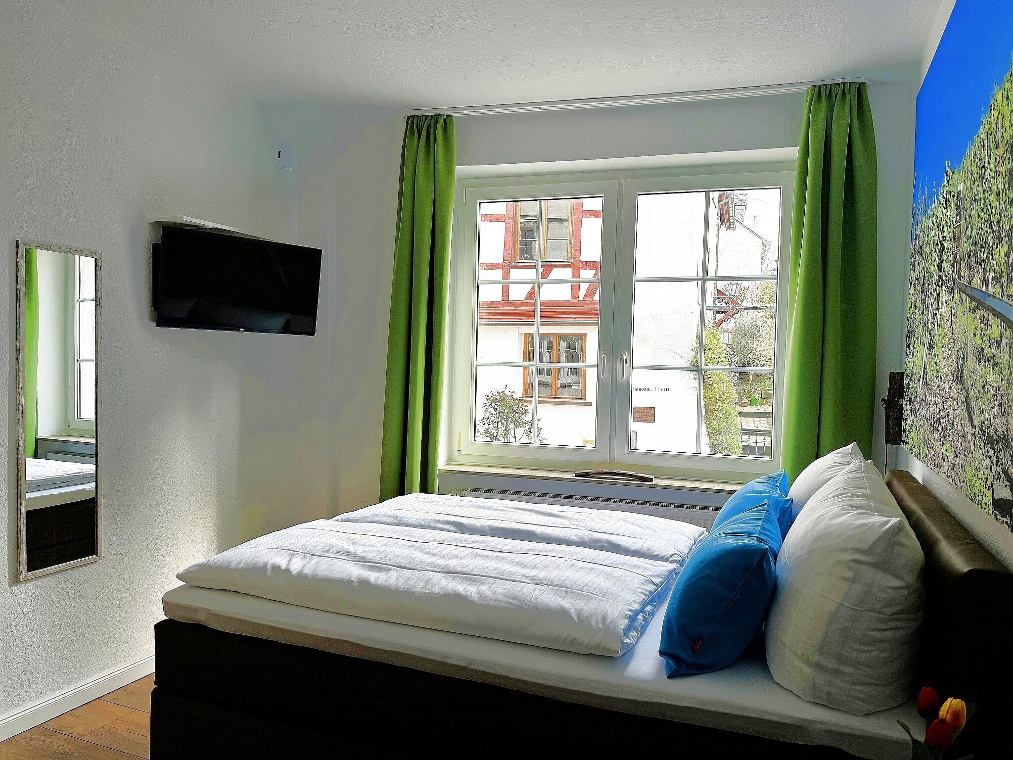 A bedroom with a bed, green curtains, a TV mounted on the wall, and a window with a view of buildings outside.