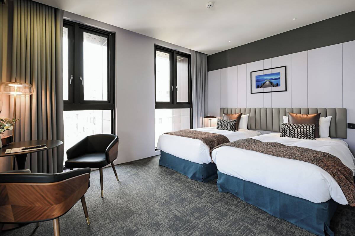 A hotel room with two beds, a desk, a chair, and large windows.