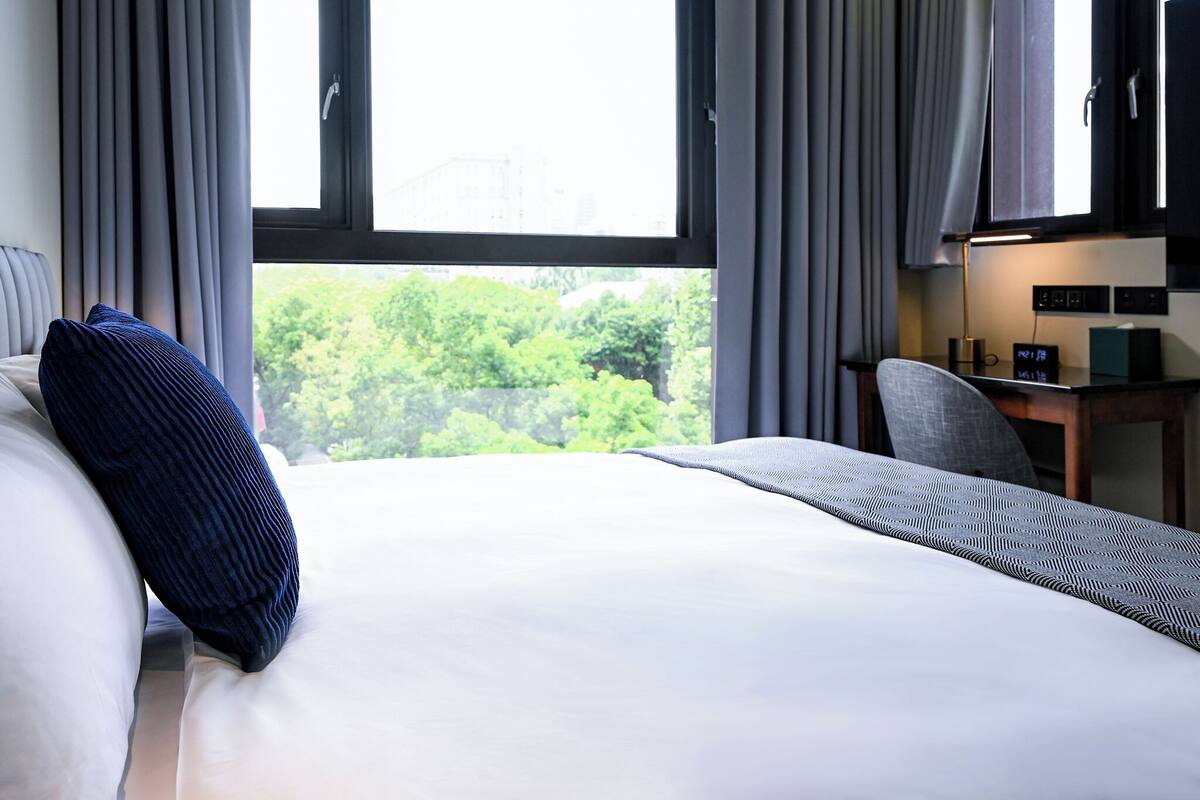 A hotel room with a bed, a desk, and a view of trees through the window.