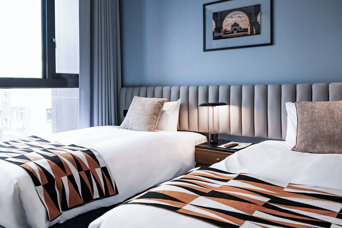 A hotel room with two beds, a nightstand, a lamp, and a framed picture on the wall.