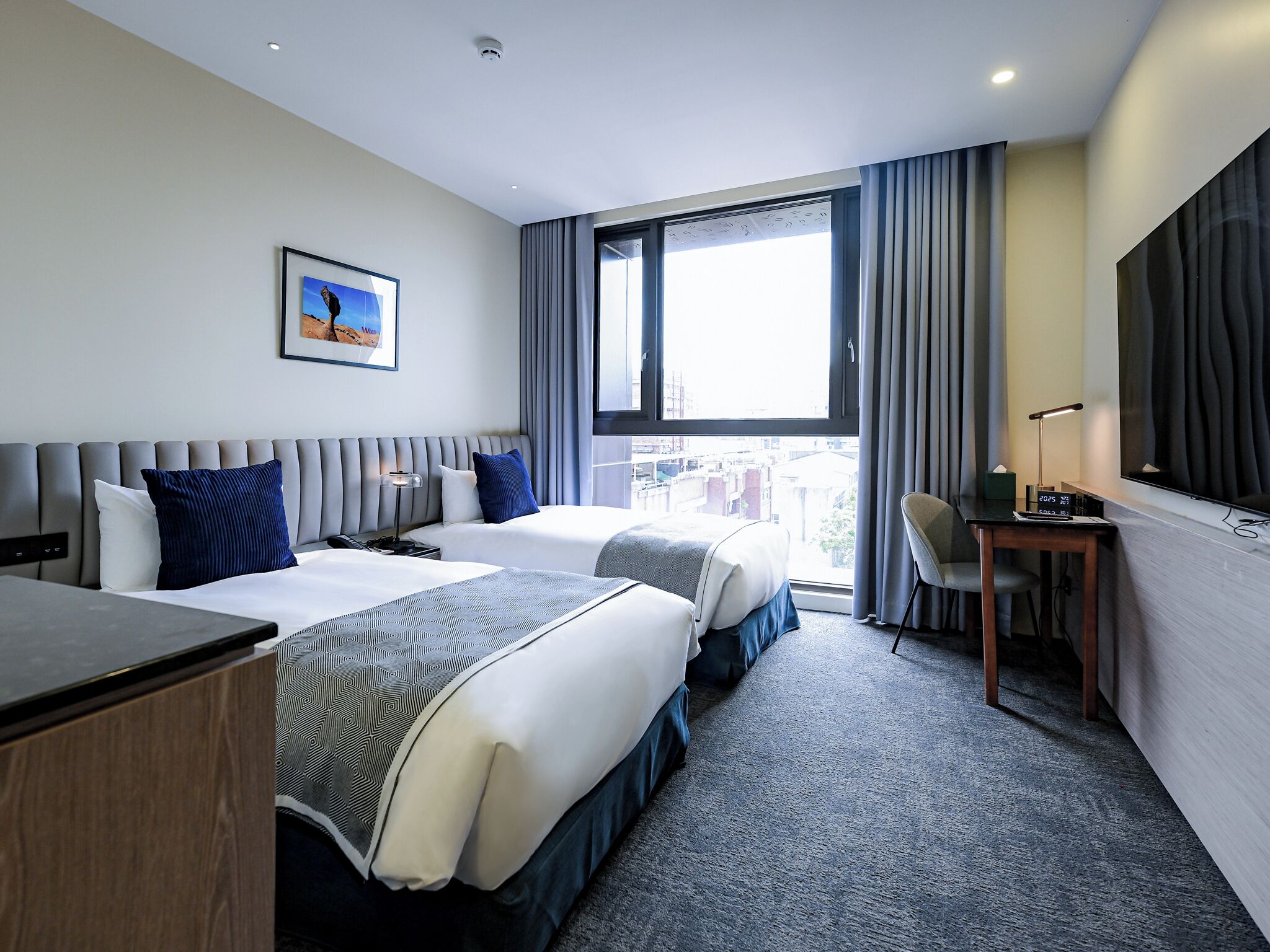 A hotel room with two beds, a desk, a chair, a TV, and a large window with curtains.