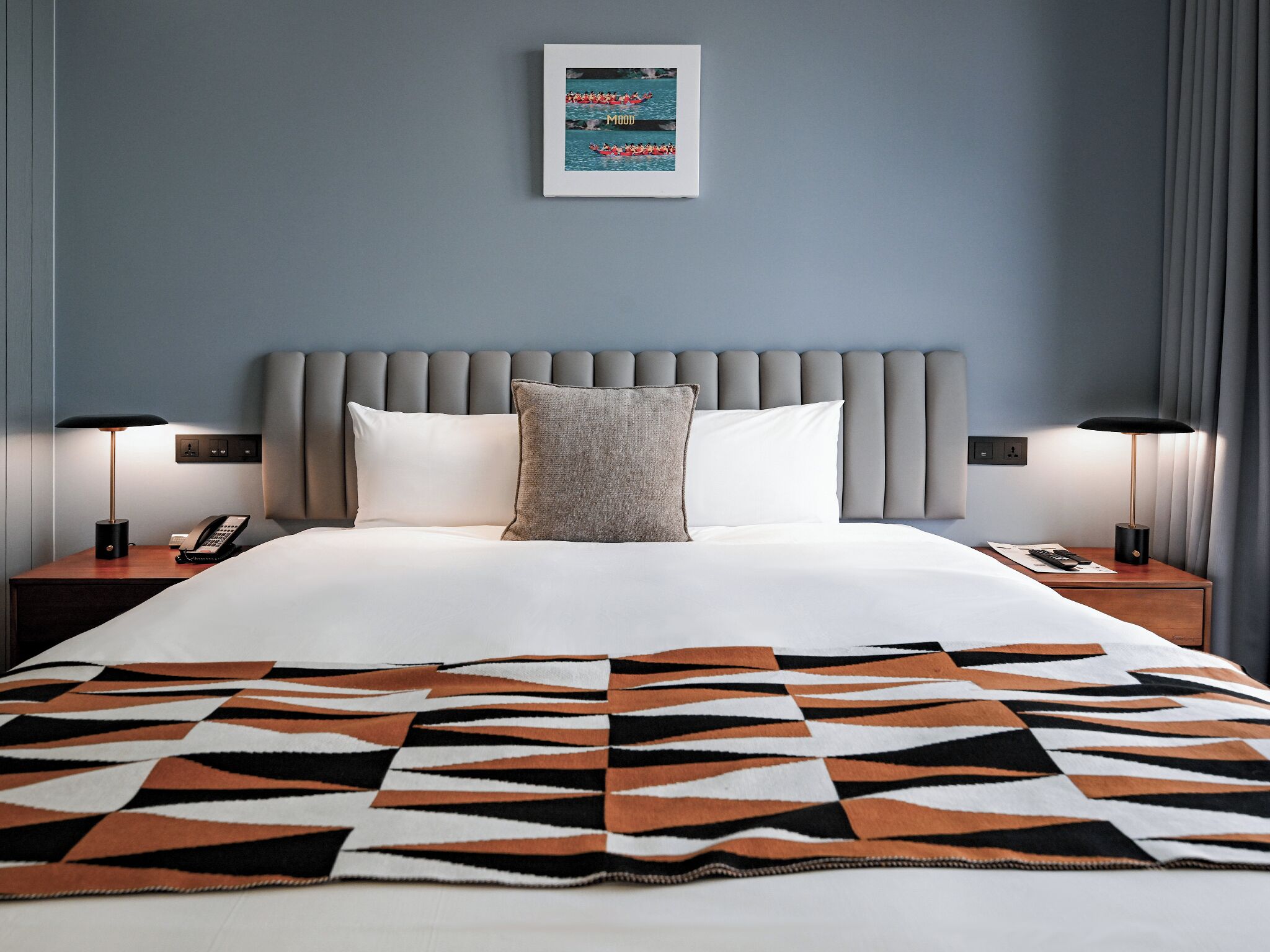 A neatly made bed with a patterned blanket, a grey upholstered headboard, bedside lamps, and a framed picture above the bed.