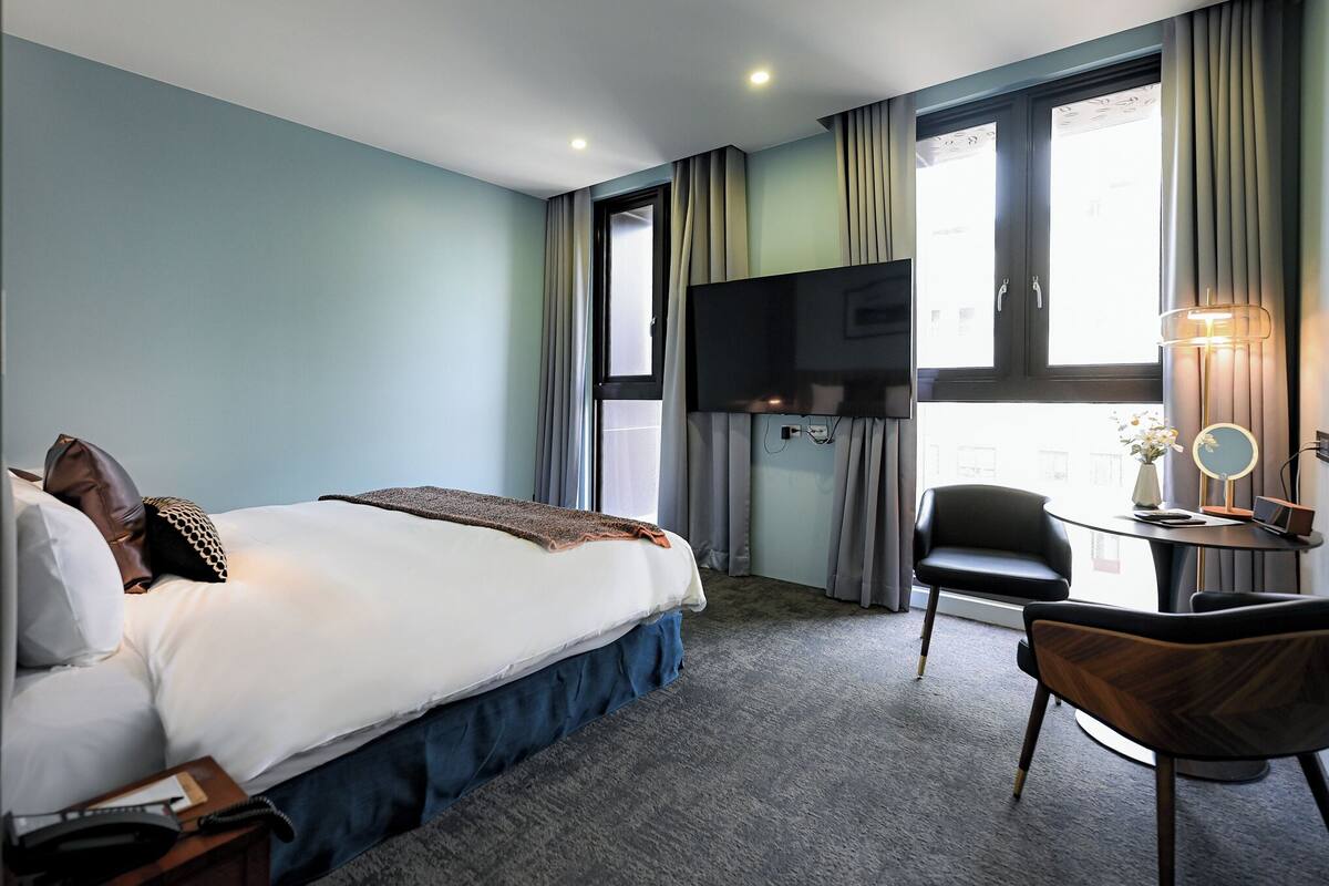 A hotel room with a bed, a desk with a chair, a TV, and a window with curtains.
