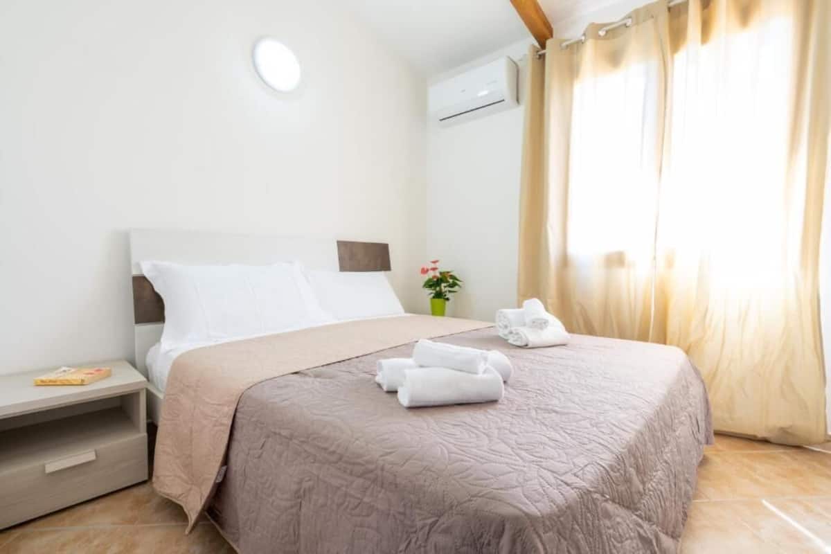 A neatly made bed with a beige quilt, a bedside table with a book, and a small potted plant.