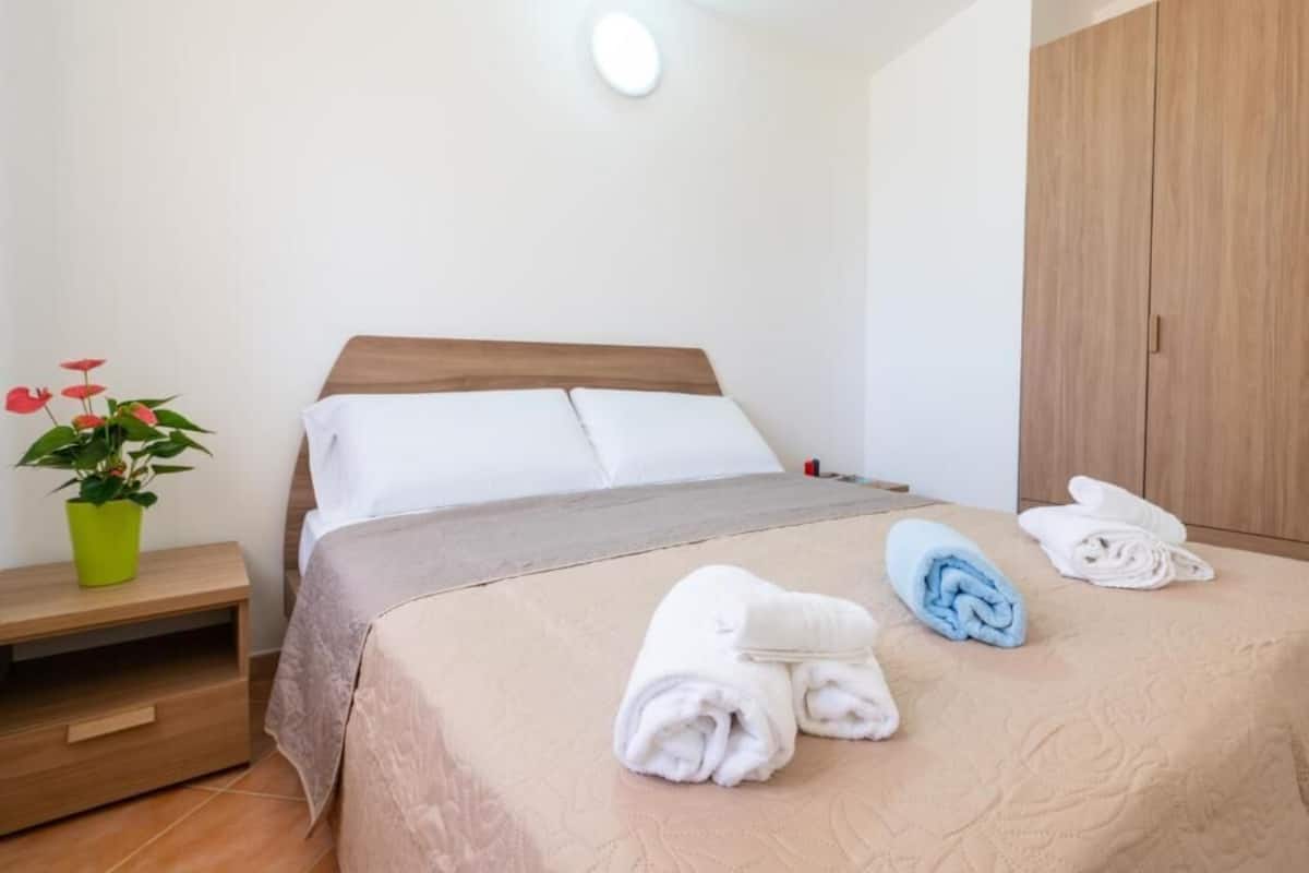 A neatly made bed with white and beige linens, a green plant on a bedside table, and a wooden wardrobe.