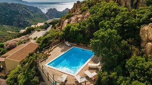 A rectangular swimming pool surrounded by white lounge chairs, set against a backdrop of lush greenery and rocky terrain with a view of the sea. - Residence La Falata (Trinità d'Agultu e Vignola)