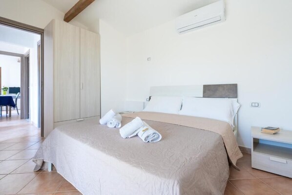 A neatly made bed with a beige bedspread and pillows, a bedside table with a book, and an air conditioning unit on the wall. - Residence La Falata (Trinità d'Agultu e Vignola)