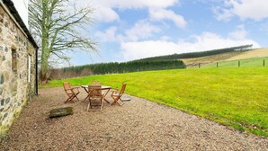 A rustic stone building with a gravel patio, wooden furniture, and a view of a green field and forest. - Westerpark Countryside Cottage Aswanley Aberdeenshire (Huntly)