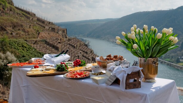 A table with various food items and a vase of white tulips, set against a scenic backdrop of a river and hills. - Ferienwohnung Kellermeister für 4 Personen in den Ferienwohnungen Bullay, Mosel (Bullay)