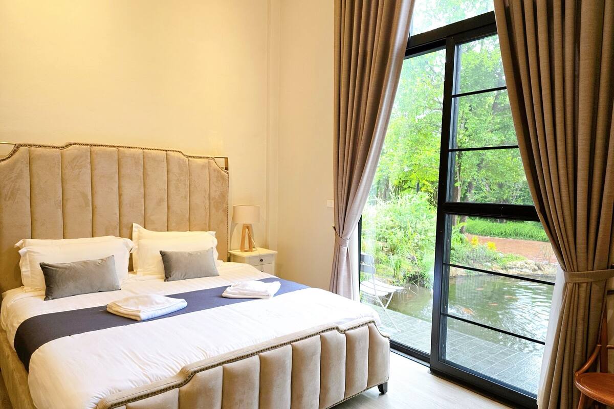 A bedroom with a large bed, a nightstand, a lamp, and a view of a garden through a window.
