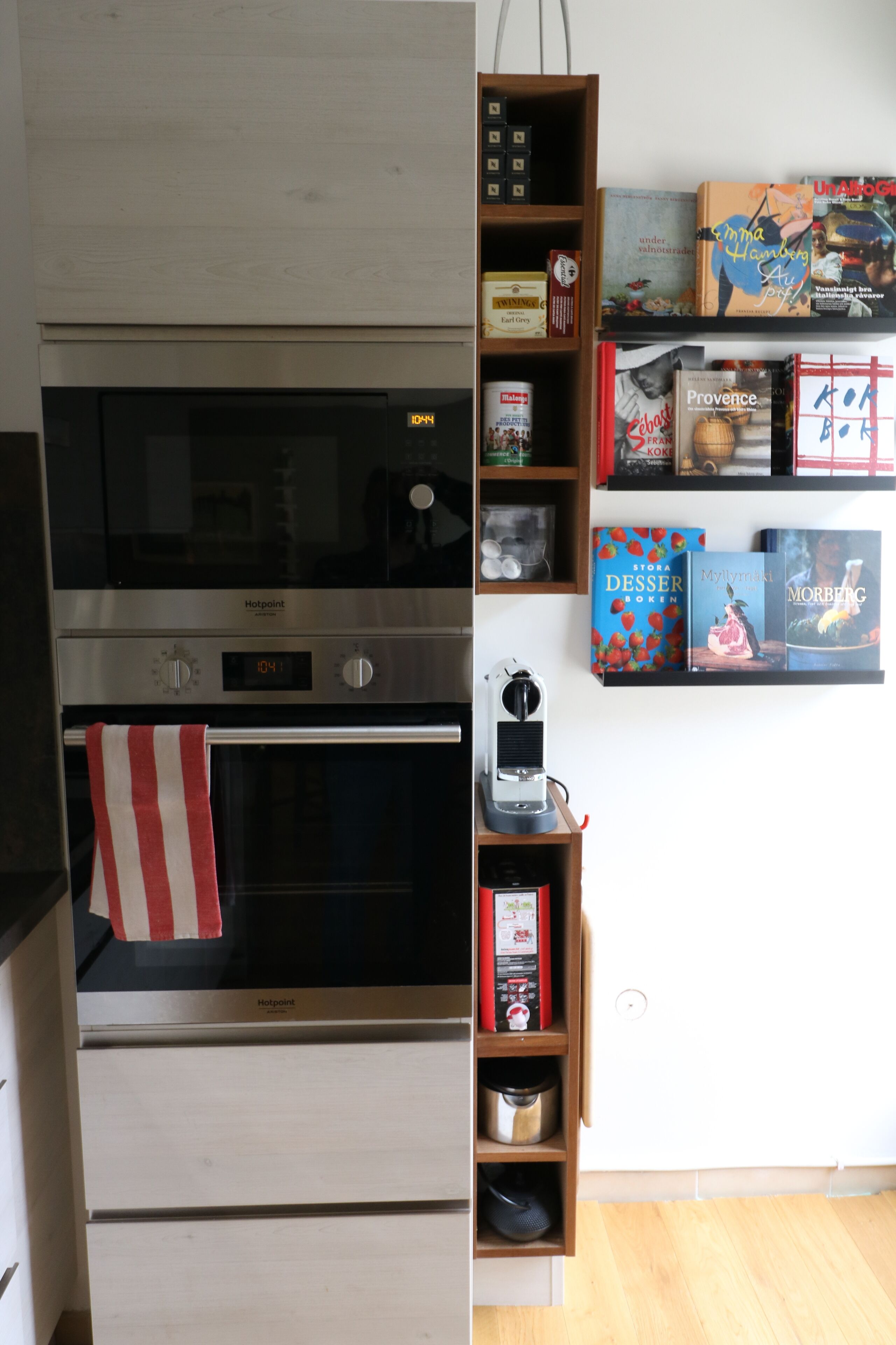 Een keuken met een ingebouwde oven, een koffieapparaat en een plank met boeken en keukengerei.