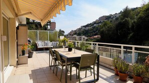 A terrace with a table and chairs, a sofa, potted plants, and a view of a hillside town. - Lovely apartment with AC and a large terrace - pool in the residence. (Nice)