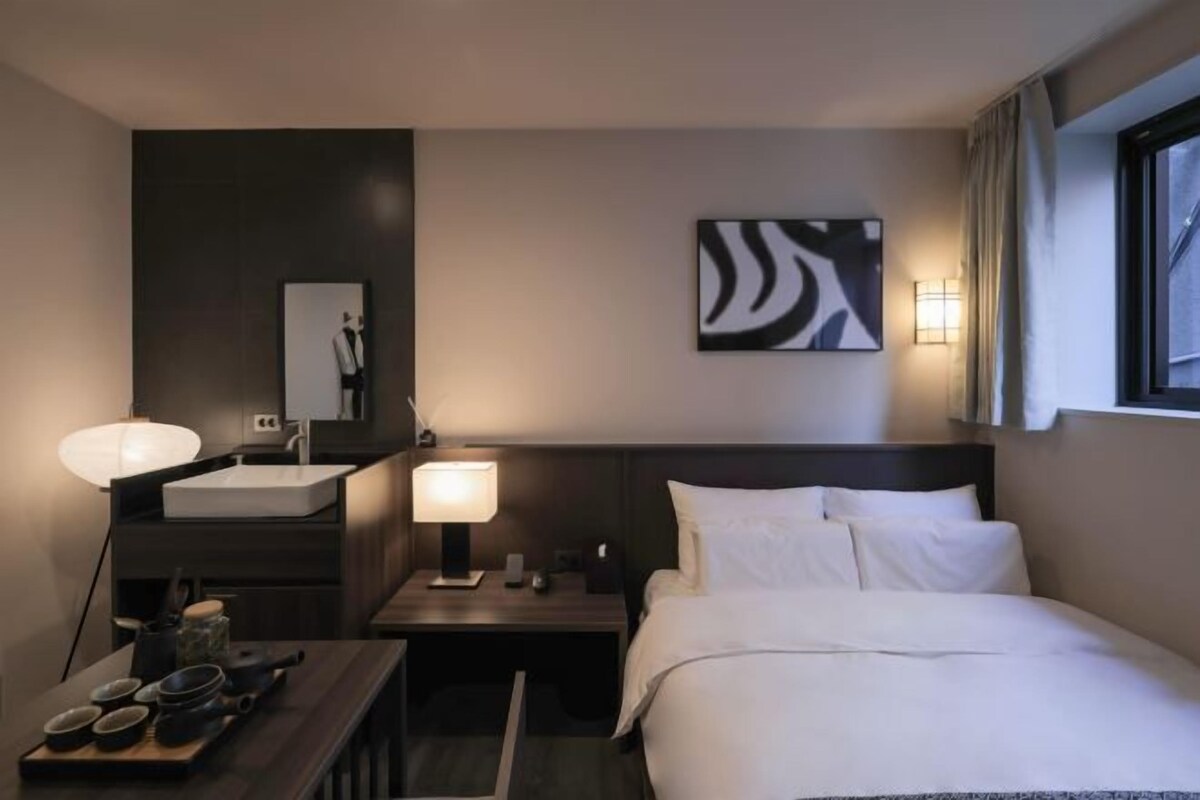 Habitación de hotel con una cama, una mesita de noche con lámpara, un espejo, una obra de arte en la pared y una ventana con cortinas.