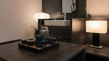 Un baño moderno con tocador de madera oscura, un lavabo blanco y un espejo encima. Hay teteras y tazas negras sobre el tocador, y una lámpara blanca a un lado.