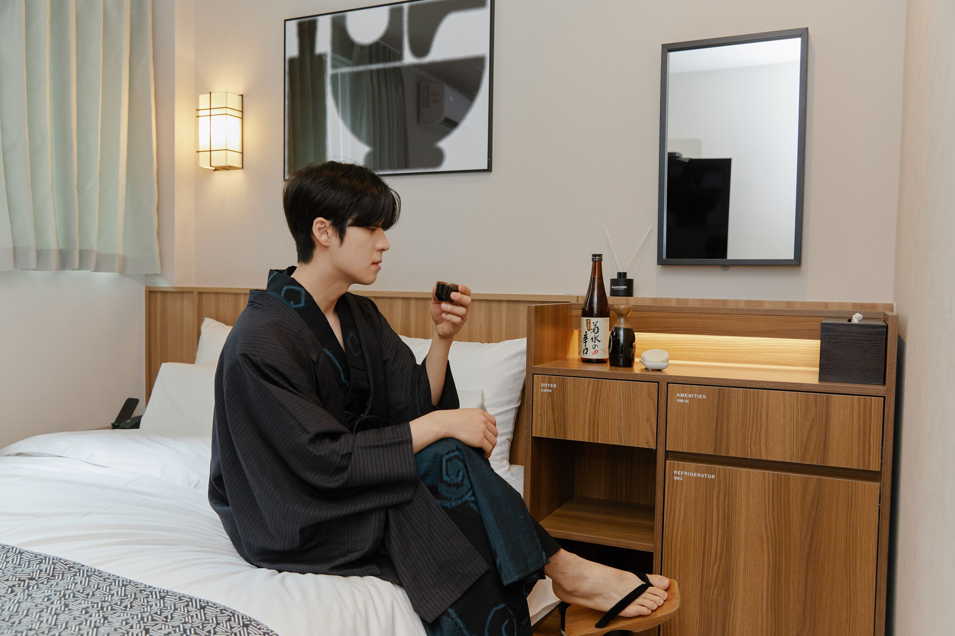 A hotel room with a bed, a bedside table with a bottle and a cup, a mirror, and a wall-mounted artwork.