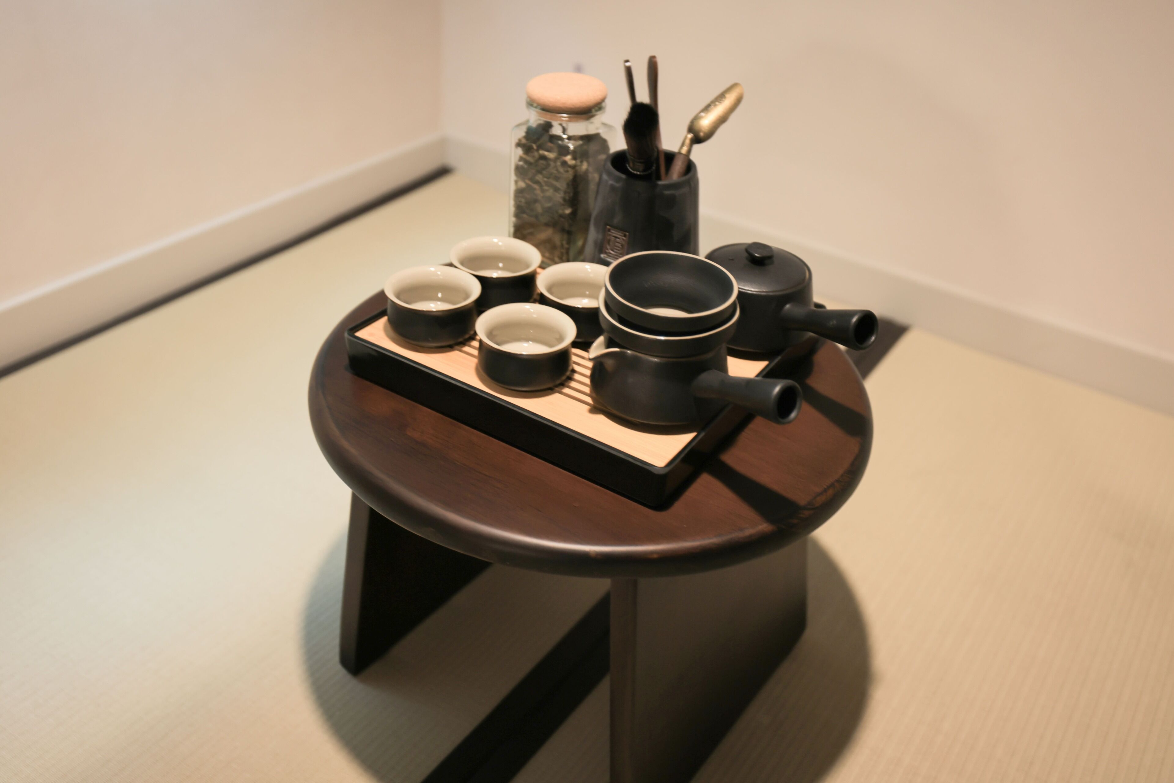 A small round table with a tray holding several black cups and a teapot, set against a plain wall.